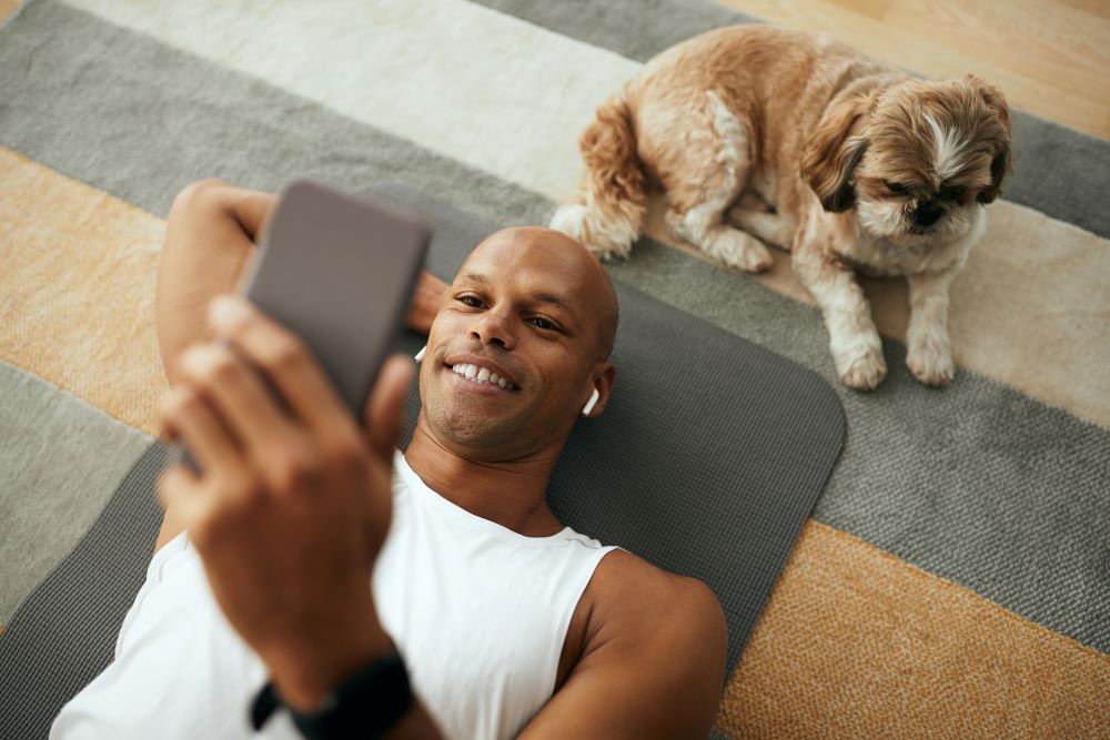 Athlete using smart phone while relaxing on floor with dog at home.